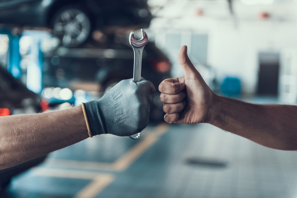 Two hands coming together in the foreground of an automotive repair bay. One hand is gloved and holding a wrench. The other is giving a thumbs up.