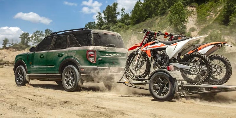 A green Bronco Sport hauling dirt bikes on a flatbed through a dusty trail backed by lightly wooded hillsides.