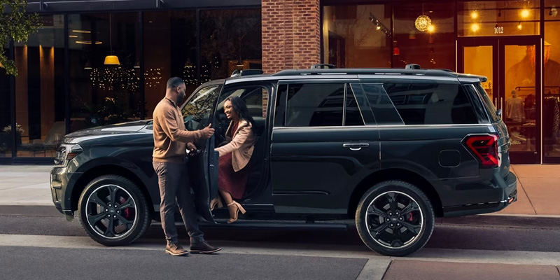 A man holding the door for a woman as she steps out of the driver's side of a black Ford Expedition, in front of a fancy restaurant.
