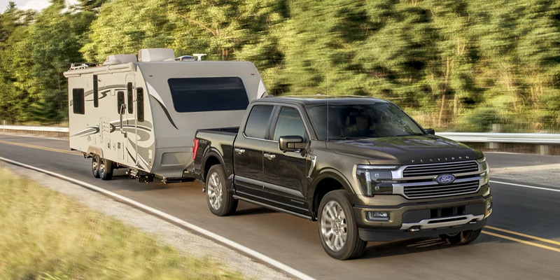 A black Ford F-150 pulling a camper trailer down a highway road, surrounded by woods and fields. 